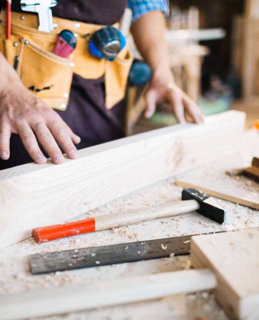 Close-up shot of male hands examining surface of wooden board, hammers, planes and saw lying on table
